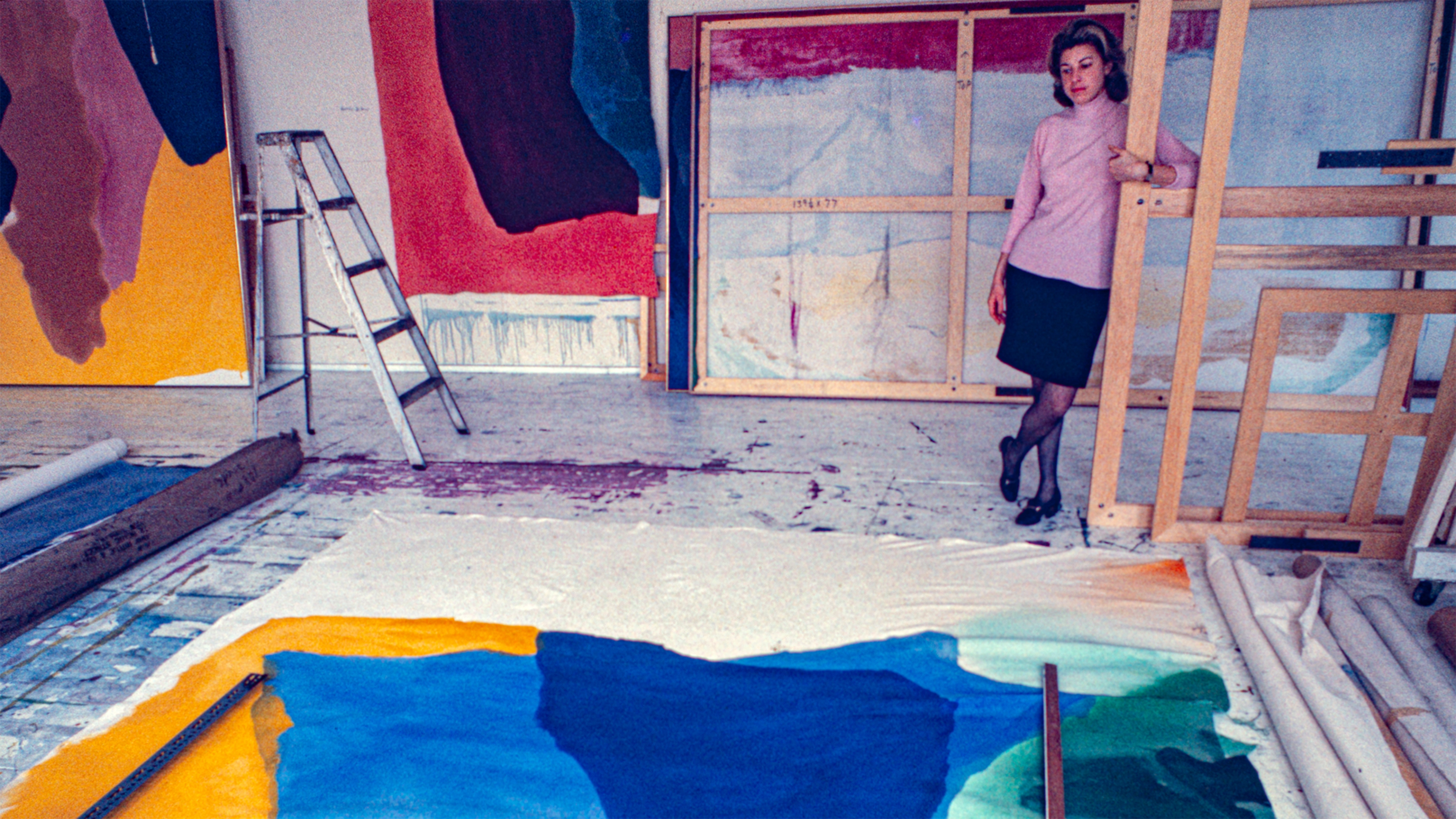 A woman stands in a colorful, paint-splattered studio surrounded by large abstract canvases.